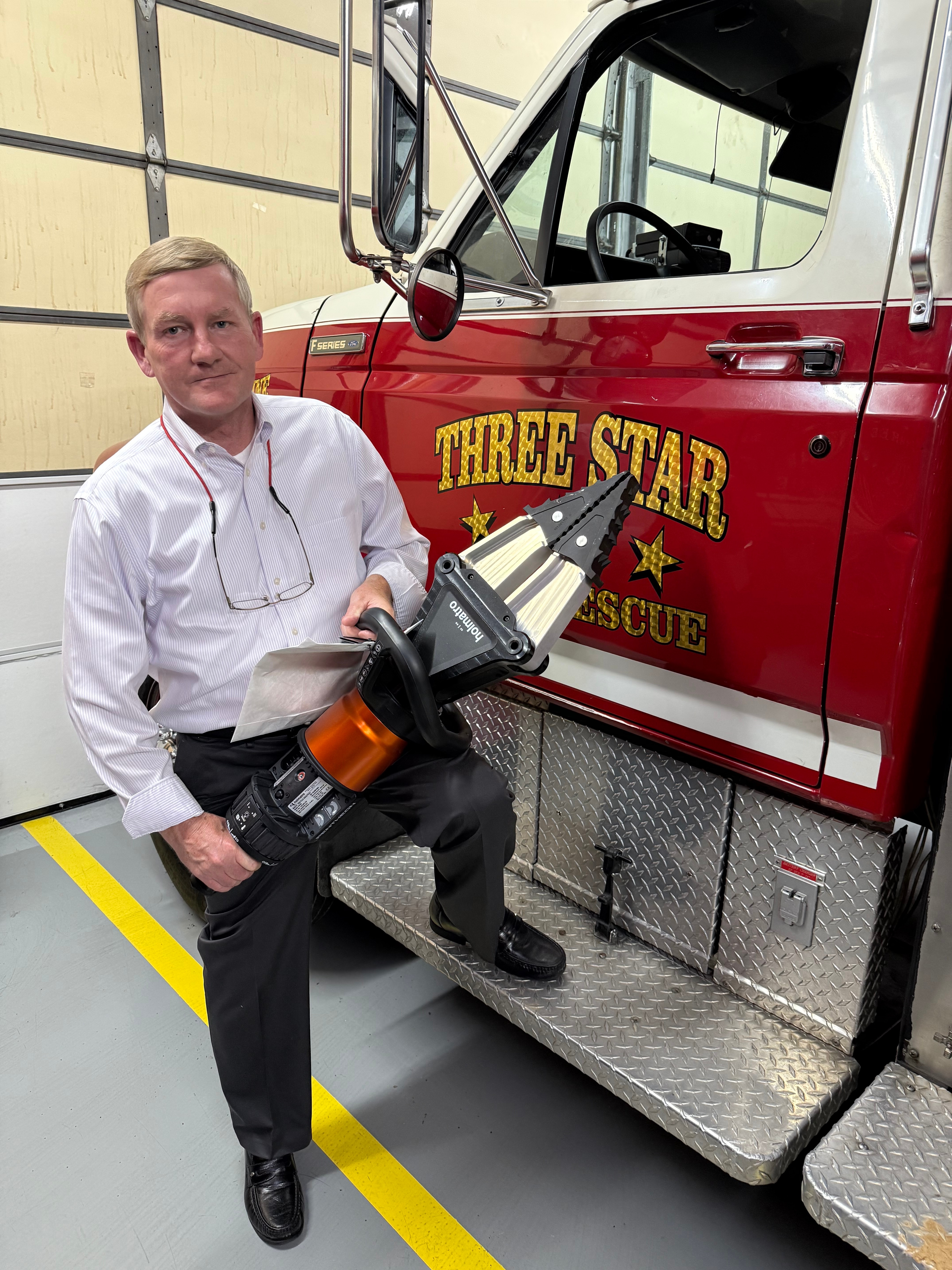 A man poses with a fore truck that says "Three Star" on the side. He has one foot up on the truck and holds a piece of extrication equipment.